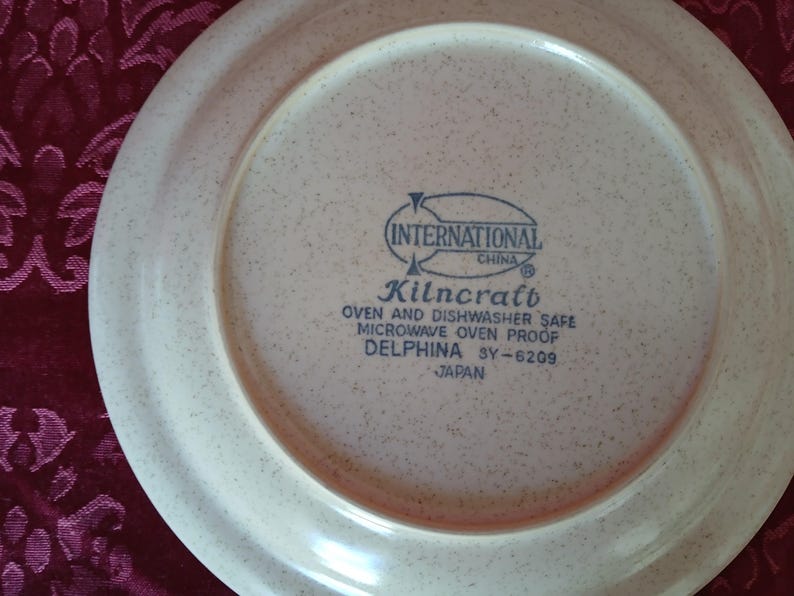 Puede incluir: Un plato de cer&aacute;mica beige con un patr&oacute;n moteado. El plato tiene un borde ancho y un &aacute;rea central con texto azul que dice "INTERNATIONAL CHINA Kilncraft OVEN AND DISHWASHER SAFE MICROWAVE OVEN PROOF DELPHINA SY-6209 JAPAN."