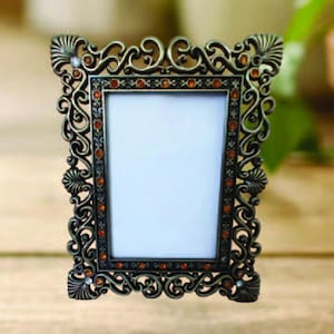 May include: Ornate bronze-colored picture frame with a rectangular opening. The frame features intricate scrollwork, shell-like accents, and small orange-colored embellishments. The frame is designed to hold a photograph or artwork, adding a touch of vintage elegance to any space.