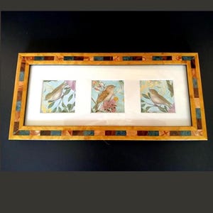 May include: A rectangular wall frame featuring a light wood finish with a mosaic border of small blue, brown, and tan tiles. The frame contains three separate square prints, each depicting a small brown bird perched among green leaves and pink flowers.