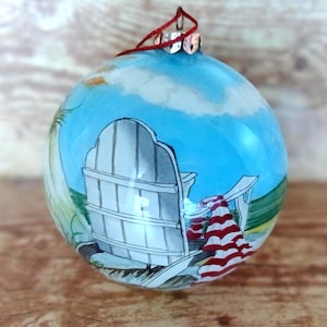 May include: A hand-painted glass Christmas ornament featuring a beach scene. The ornament depicts a white Adirondack chair on a sandy shore with a red and white striped towel. The background shows a blue sky and ocean.