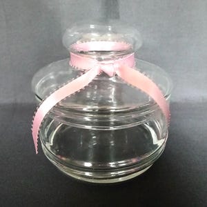May include: A clear glass jar with a round body and a lid. A pink ribbon with a scalloped edge is tied around the jar. The jar has a small, round top. The jar is likely used for storage or decoration.
