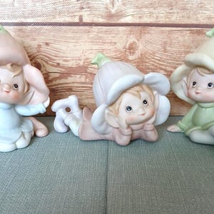 May include: Three ceramic figurines of flower fairies in pastel colors. One is kneeling in a light blue outfit, another is lying down in a pink outfit, and the third is sitting in a green outfit. Each has a flower-shaped hat.