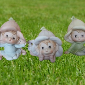 May include: Three ceramic figurines of flower fairies on a grassy background. Each fairy has a flower-shaped hat and is dressed in pastel colors: light blue, pink, and green. The fairies are in different poses.