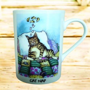 May include: White ceramic mug featuring a whimsical illustration of a tabby cat napping on a stack of colorful pillows. The cat dreams of fish, with the words "CAT NAP" at the base. The mug has a curved handle.
