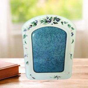 May include: A white, arched photo frame with a blue textured insert. The frame is decorated with green leaves and blue berries. The frame is approximately 5x7 inches.