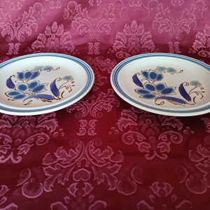 Puede incluir: Dos platos ovalados de cer&aacute;mica. Cada plato presenta una base blanca con un dise&ntilde;o floral azul y un borde azul y blanco. El dise&ntilde;o floral incluye flores azules y tallos marrones. Los platos est&aacute;n sobre un fondo rojo estampado. Cada plato mide unos 25 cm.