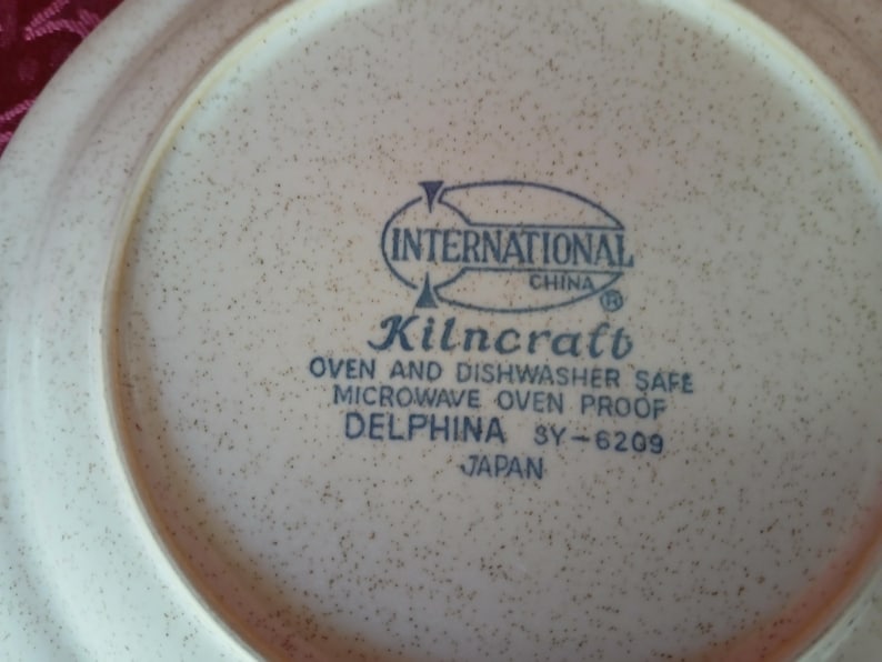Puede incluir: Primer plano de un plato de cer&aacute;mica beige con un patr&oacute;n moteado. El plato presenta el texto "INTERNATIONAL CHINA Kilncraft OVEN AND DISHWASHER SAFE MICROWAVE OVEN PROOF DELPHINA SY-6209 JAPAN".