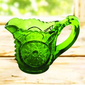 May include: A green glass creamer with a decorative starburst pattern on the front. The creamer has a ruffled rim and a curved handle. The glass is a vibrant shade of green, and the creamer is sitting on a wooden surface.