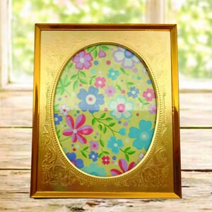 May include: A rectangular gold-colored picture frame with an oval opening. The frame has ornate detailing and holds a floral print with pink, blue, and white flowers on a yellow-green background. The frame is displayed on a wooden surface.