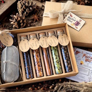 May include: A gift set for mulled wine, featuring a box with a tea infuser, glass tubes filled with spices, and a gift box tied with twine. The tubes contain cloves, orange peel, cinnamon sticks, and more. A tag reads "Mulled Wine".