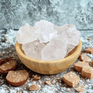 Puede incluir: Un cuenco de madera lleno de grandes trozos cristalinos translúcidos. El cuenco está sobre una superficie salpicada de polvo blanco y pequeños trozos marrones de forma irregular. El fondo es una superficie gris texturizada.