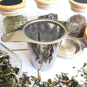 May include: A stainless steel tea infuser with a handle and a fine mesh strainer. The infuser is sitting on a white surface with loose leaf tea scattered around it.