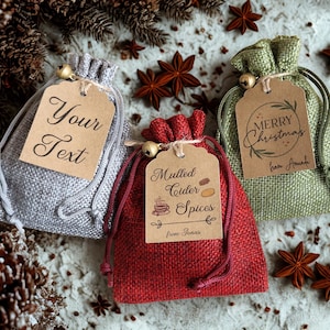 May include: Three small burlap drawstring pouches in silver, red, and green. Each bag features a brown paper tag tied with twine and a small bell. The tags display the text Your Text, Mulled Cider Spices from Jonas, and Merry Christmas from Amanda.