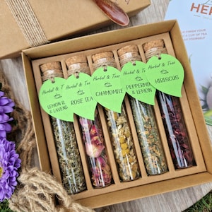 Könnte beinhalten: Ein Set aus sechs Glasröhrchen gefüllt mit getrockneten Kräutern, jedes mit einem grünen, blattartigen Etikett, das die Art des Krauts kennzeichnet: Zitrone & Lavendel, Rosen Tee, Kamille, Pfefferminze & Zitrone und Hibiskus. Die Röhrchen sind in einer Holzkiste mit braunem Deckel angeordnet.