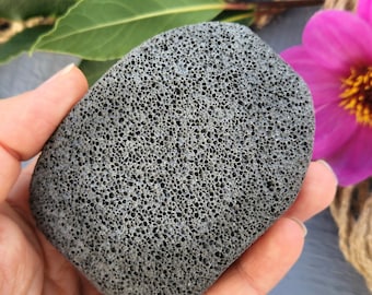 Puede incluir: Una piedra pómez gris con una textura porosa, sostenida en una mano. La piedra tiene forma ovalada y parece ser para exfoliar. El fondo incluye hojas verdes y una flor rosa.