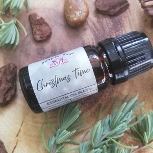May include: A dark glass essential oil bottle with a black cap, labeled "Christmas Time Essential Oil Blend." The label features the text in a script font. The bottle is surrounded by greenery, bark, and stones on a wooden surface.