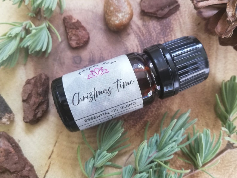 May include: A dark glass essential oil bottle with a black cap, labeled "Christmas Time Essential Oil Blend." The label features the text in a script font. The bottle is surrounded by greenery, bark, and stones on a wooden surface.