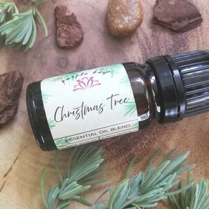 May include: A small, dark amber glass bottle with a black cap, labeled "Christmas Tree Essential Oil Blend." The label features green accents and the text "100% pure oil." The bottle is surrounded by sprigs of greenery and small pieces of bark.