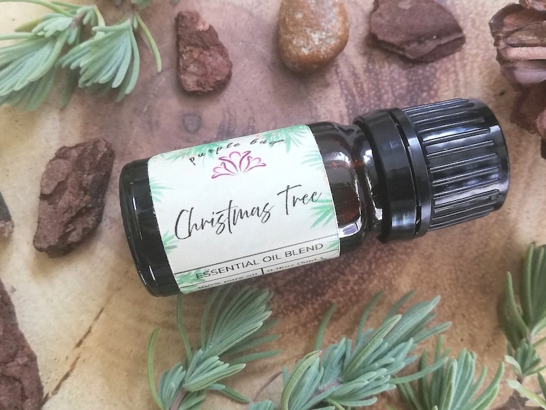 May include: A small, dark amber glass bottle with a black cap, labeled "Christmas Tree Essential Oil Blend." The label features green accents and the text "100% pure oil." The bottle is surrounded by sprigs of greenery and small pieces of bark.