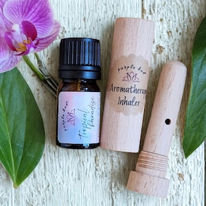 Puede incluir: Juego de aromaterapia con una botella de vidrio marrón, tapa negra y etiqueta "Tropical Paradise". Incluye un inhalador de aromaterapia de madera y un estuche. También hay una flor de orquídea púrpura y hojas verdes.