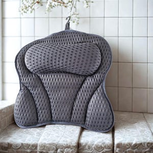 Puede incluir: Una almohada de baño gris con una superficie texturizada y un reposacabezas integrado. La almohada tiene una forma curva diseñada para sostener la espalda y el cuello. Incluye un lazo para colgar. El fondo presenta azulejos blancos y una ventana.