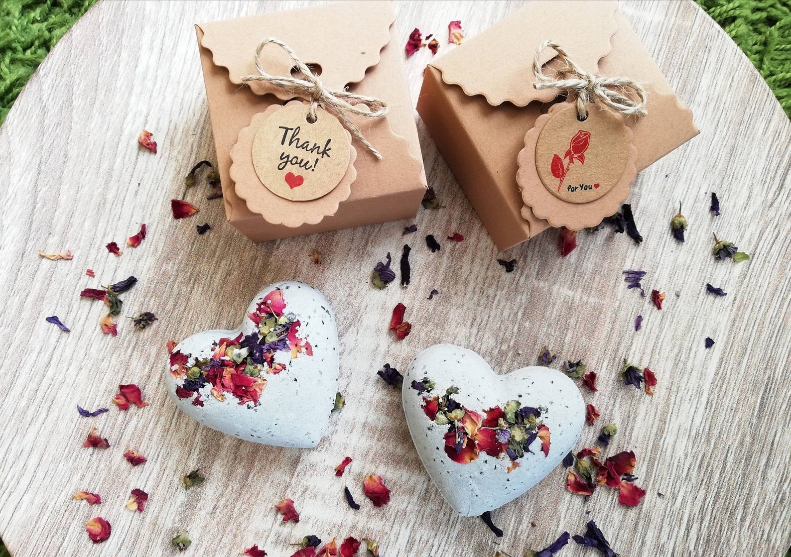 Two gift boxes with twine bows and heart-shaped tags sit on a wooden surface. Nearby, two heart-shaped soaps with colorful dried flowers.