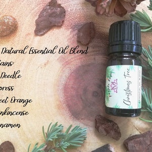 May include: A small, dark glass bottle of "Christmas Tree" essential oil blend. The label lists ingredients: fir needle, cypress, sweet orange, frankincense, and cinnamon. The bottle sits on a wooden surface with sprigs of greenery and bark.