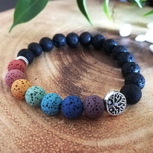 Puede incluir: Pulsera de piedra de lava arcoíris con un dije de árbol de la vida plateado. La pulsera está hecha de cuentas de piedra de lava negra con un arcoíris de colores en el centro.