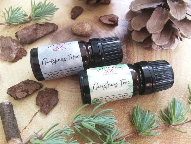 May include: Two small essential oil bottles with black caps and labels that read "Christmas Time" and "Christmas Tree." The bottles are surrounded by pine needles, bark, and a pine cone on a wooden surface.