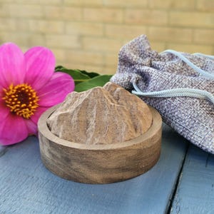 May include: A wooden coaster with a mountain design sits on a blue surface next to a pink flower and a grey drawstring bag. The coaster is round with a carved mountain range on top. The flower has a yellow centre.