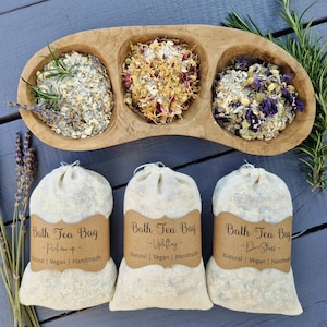 May include: Three white muslin bags filled with dried herbs and flowers for bath tea. The bags are labelled "Bath Tea Bag" with different scents: "Pick me up", "Uplifting", and "De-Stress". The bags are arranged on a wooden surface with a bowl of dried herbs and flowers in the background.