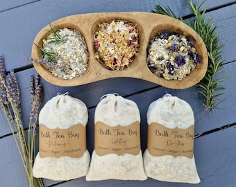 Puede incluir: Una fuente de madera con tres secciones llenas de hierbas y flores secas. Tres bolsas de té de baño beige con etiquetas marrones están al frente. Las etiquetas dicen "Bath Tea Bag" con diferentes lemas. Ramitas de lavanda y romero están a un lado.