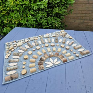 Op de afbeelding: Een lichtblauw tapijt met een decoratief ontwerp gemaakt van houten schijfjes, kleine stenen en houten uitsparingen. De opstelling omvat een halfrond patroon en een kleine houten voetafdruk uitsparing. Het tapijt ligt op een blauw houten oppervlak.