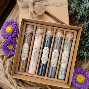 Puede incluir: Un juego de regalo de colección de sal gourmet en una caja marrón. El juego incluye seis viales de vidrio llenos de diferentes tipos de sal, etiquetados como "Smoked Salt", "Himalayan Salt", "Black Lava Salt", "Kala Namak" y "Herb Salt". La caja está atada con hilo y una etiqueta.