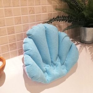 Puede incluir: Una almohada de baño azul claro en forma de concha marina descansa sobre el borde de una bañera blanca. Un cuenco de madera lleno de sales de baño y una pequeña cuchara de madera están cerca. Un helecho en maceta está en el fondo.