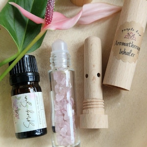 Puede incluir: Un conjunto de productos de aromaterapia que incluye un frasco de vidrio con rodillo lleno de cristales rosas, una pequeña botella de vidrio marrón etiquetada como "Spring Sun Essential Oil Blend" y un inhalador de aromaterapia de madera con una etiqueta que dice "Purple Bloom Aromatherapy Inhaler".