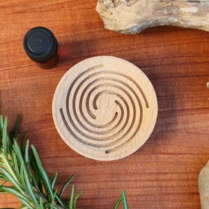 May include: A circular wooden aromatherapy diffuser with a spiral pattern, alongside a small brown bottle with a black cap. Fresh green rosemary sprigs are on the left. The items are on a brown wooden surface.