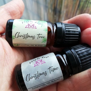May include: Two small brown glass essential oil bottles with black caps are held in a hand. One bottle is labeled "Christmas Tree" and the other "Christmas Time". Both labels feature the "purple bug" logo and the words "essential oil blend".