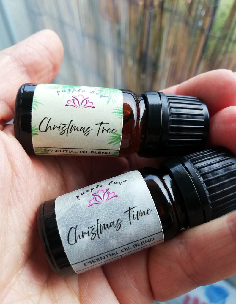 May include: Two small brown glass essential oil bottles with black caps are held in a hand. One bottle is labeled "Christmas Tree" and the other "Christmas Time". Both labels feature the "purple bug" logo and the words "essential oil blend".