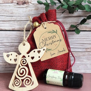 May include: A light-coloured wooden angel ornament with heart cutouts and spiral patterns, a red burlap drawstring pouch, a brown paper gift tag with the text Merry Christmas from Amanda, and a small amber glass bottle of Christmas Tree oil.