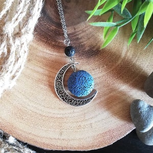 Può includere: Collana argentata con un ciondolo a forma di mezzaluna dal design intricato, che contiene una sfera blu. La catena presenta anche una perla nera e una sfera blu. La collana è presentata su una superficie di legno.