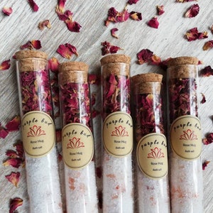 Op de afbeelding: Vijf kleine glazen buisjes gevuld met witte badzout en gedroogde rozenblaadjes. Elke buis heeft een kurkstop en een bruin papieren etiket met de tekst "purple bud" en "Rose Hug Bath Salt".