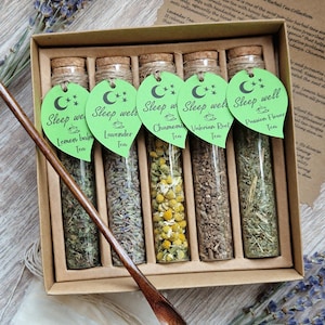 May include: A gift set featuring five glass vials of herbal tea, each with a green tag and cork stopper. The vials contain dried herbs, and a wooden spoon is placed in front. The tags read "Sleep well" with tea names.