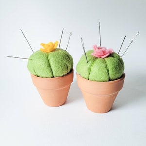 May include: Two green felt cactus pincushions in terracotta pots. One cactus has a yellow felt flower and the other has a pink felt flower. Both have metal pins sticking out.