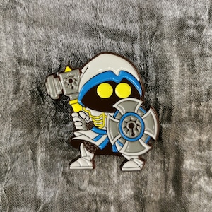 Könnte beinhalten: Ein Emaille-Pin mit einer Cartoon-Figur in einem weiß-blauen Kapuzen-Outfit, die einen gelben Hammer und einen grauen Schild hält. Die Figur hat gelbe Augen, vor einem strukturierten grauen Hintergrund.