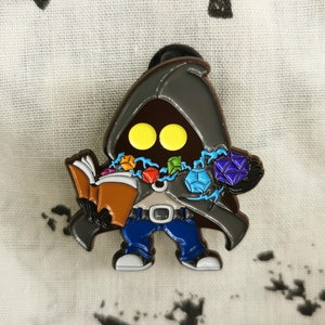 Op de afbeelding: Een cartoon-stijl emaille pin met een figuur met gele ogen, die een boek en een handvol kleurrijke edelstenen vasthoudt. De figuur draagt een blauwe spijkerbroek en een grijze cape. De pin staat tegen een witte stoffen achtergrond.