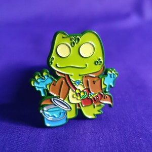 Puede incluir: Un pin de esmalte de rana verde con un abrigo marrón, sosteniendo un cubo y un pincel azules. La rana tiene grandes ojos amarillos y una bandolera roja. El pin está sobre un fondo morado.