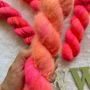 Puede incluir: Primer plano de madejas de hilo rosa y naranja vibrantes. El hilo es suave y esponjoso, con un degradado de colores que va del rosa brillante al naranja cálido. Las madejas están retorcidas y listas para proyectos de manualidades.