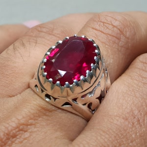 Peut inclure: Une bague en argent avec une pierre précieuse rubis rouge foncé de forme ovale sertie dans un design orné et détaillé.