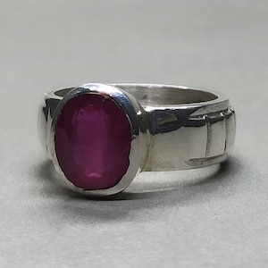 May include: A silver ring featuring a deep pink oval gemstone set in a bezel setting. The band is textured with a repeating pattern.